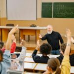 Professor engaging with students in a lively university classroom discussion as students raise hands.
